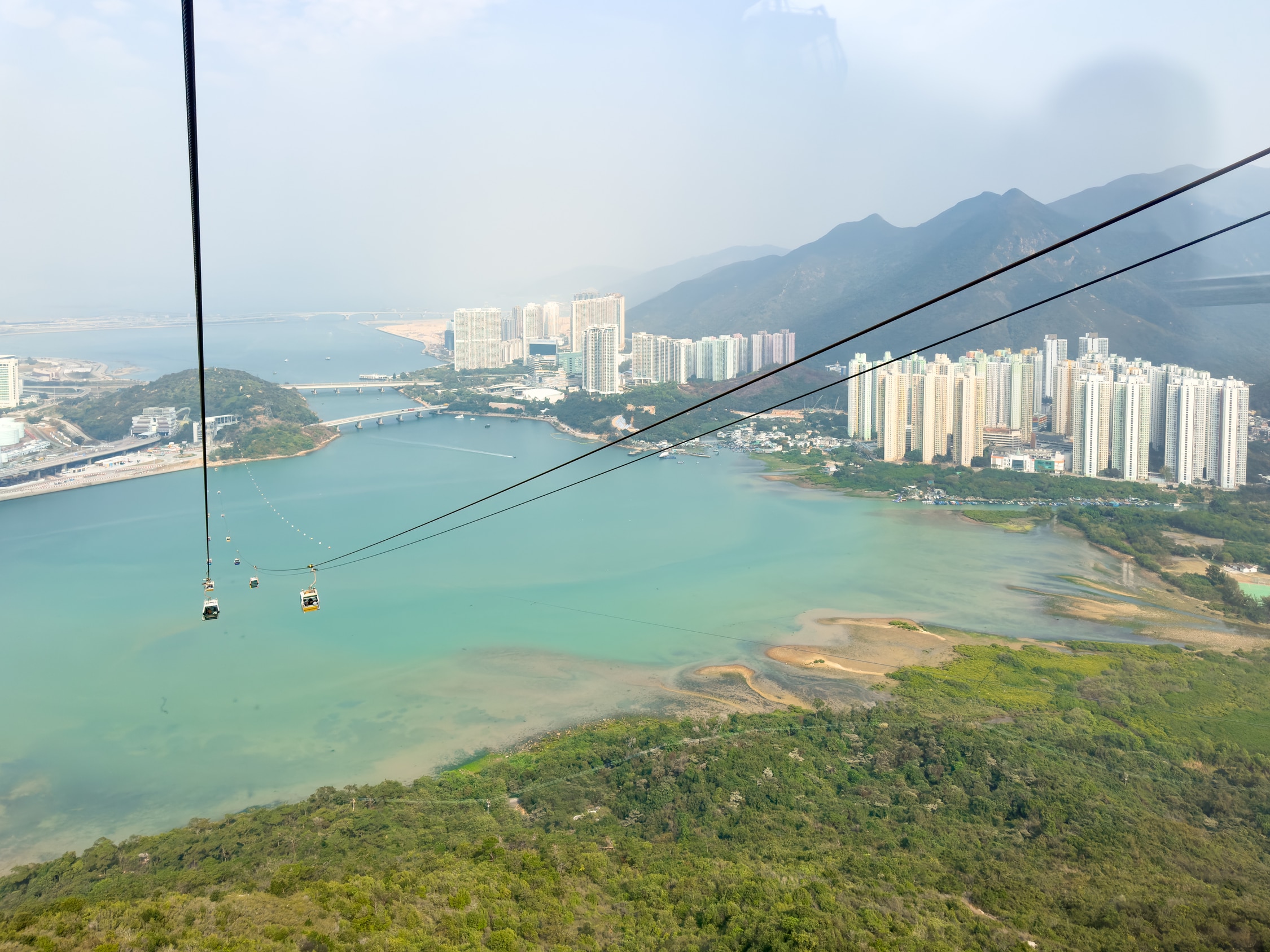 Blick aus der Seilbahn entlang der Kabel einen Berg hinunter. Die Seilbahn ist über das Meer bis zu einem Berg auf einer anderen Insel gespannt. Auf der Rechten Seite sieht man wieder Berge und Hochhäuser.