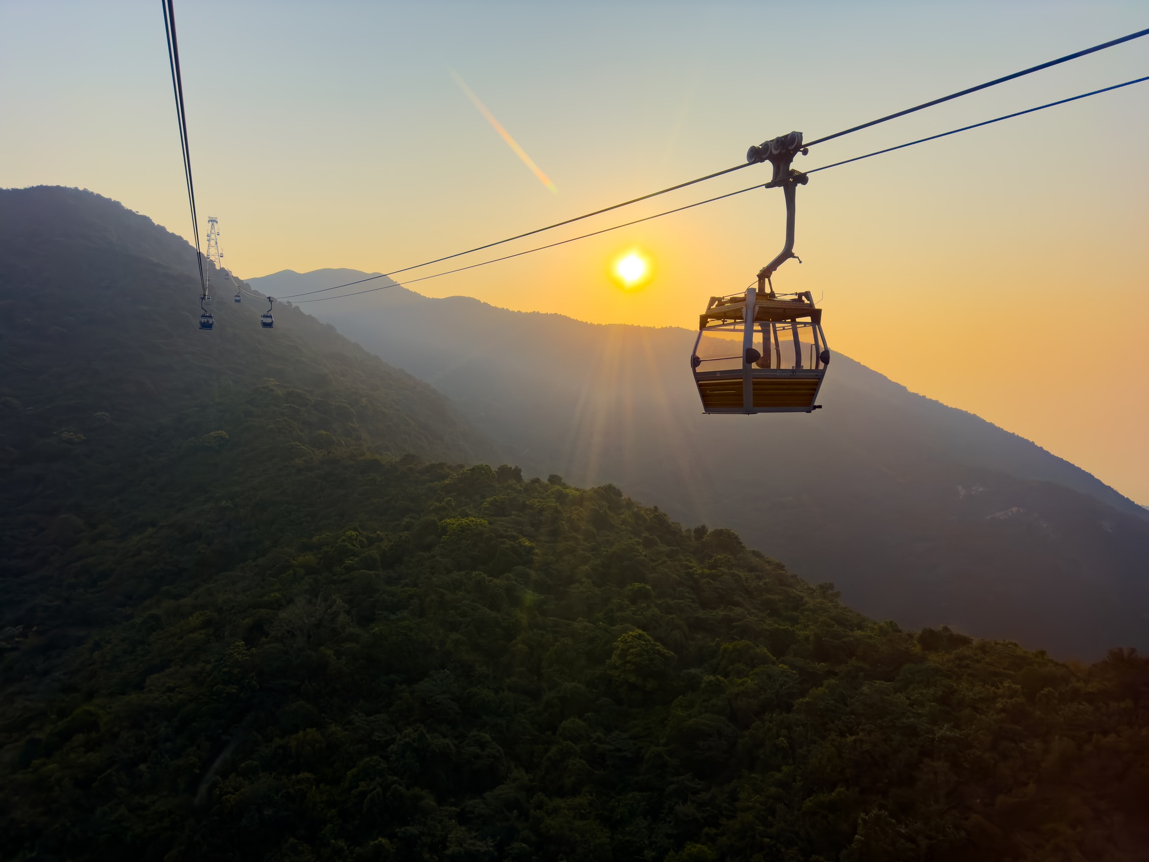 Siluette einer Gondel der Seilbahn im entgegen Leuchtenden Sonnenuntergangslicht. Im Hintergrund Berge in leichtem Nebel.